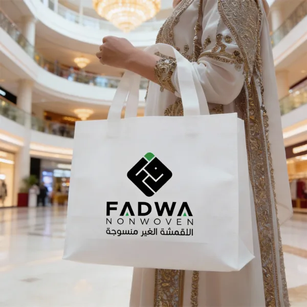 A close-up shot of a white non-woven box bag with a black and green corporate Fadwa logo, held by a person in elegant white attire inside a luxury building.