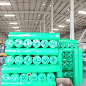 A close-up view of a large green-wrapped roll of non-woven PP agricultural crop cover fabric stacked inside a manufacturing warehouse.